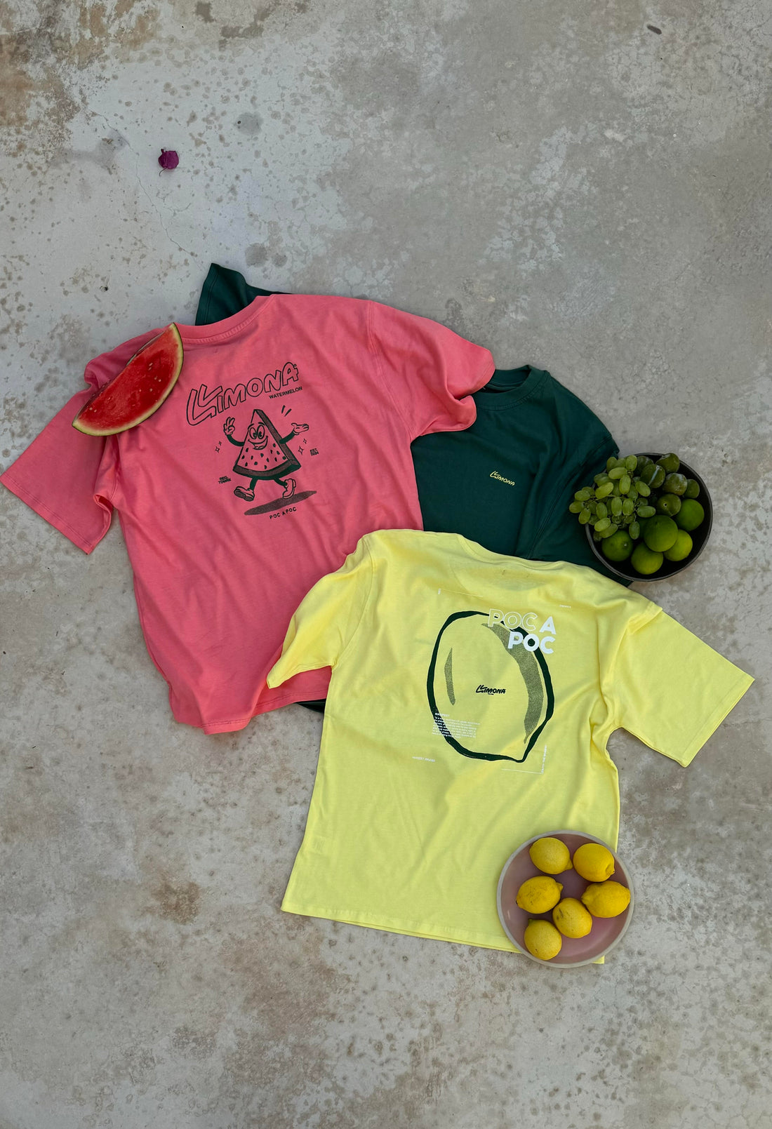 Camisetas coloridas en rosa, verde y amarillo con gráficos y logotipos de frutas, exhibidas sobre una superficie de concreto junto a sandía, uvas y limones. Perfectas para moda veraniega vibrante y estilo casual.